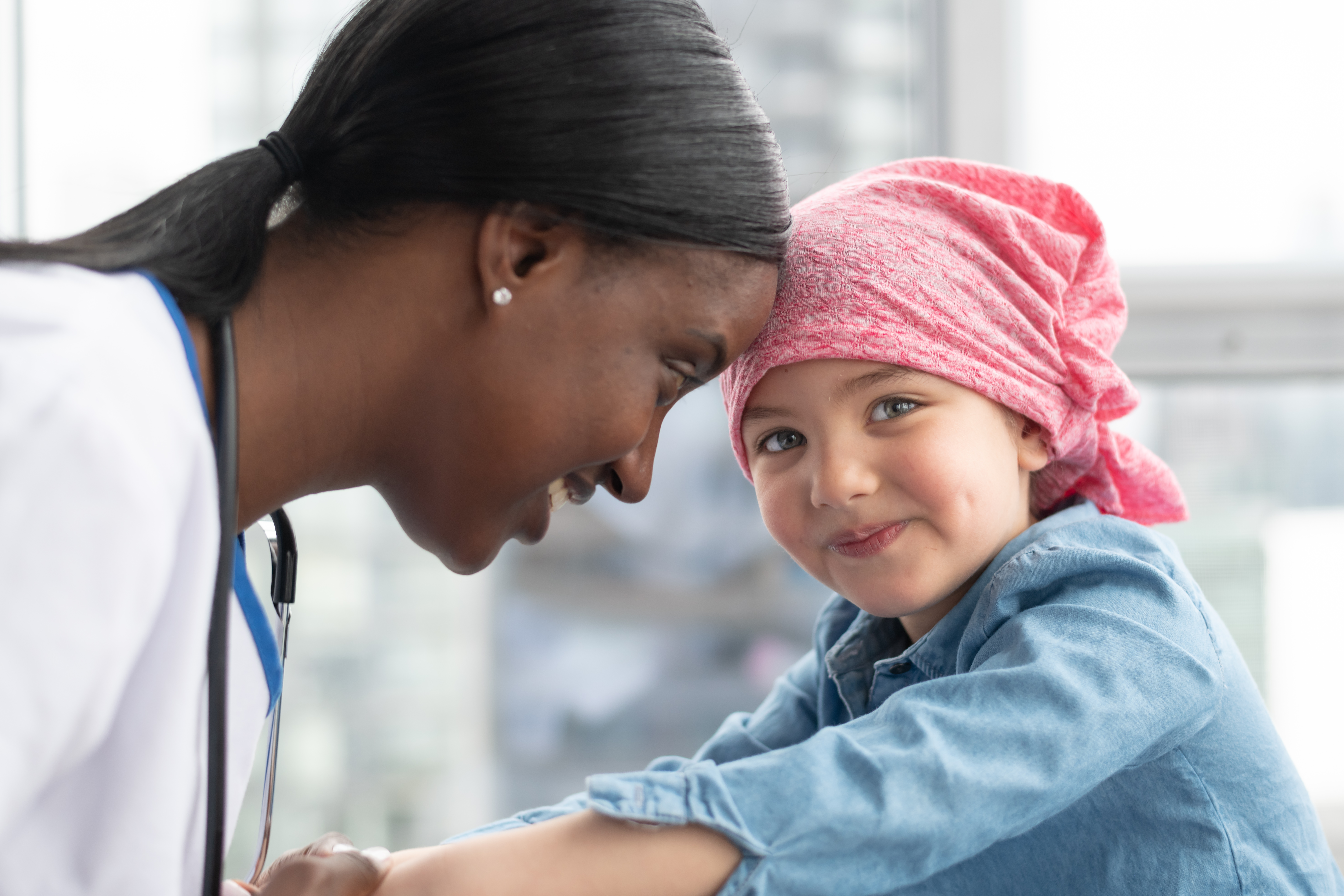A cute elementary age girl with cancer is wearing a pink scarf on her head. She is at a medical appointment. The female doctor of African descent is holding the child's hands, providing comfort and support. The child is smiling at the camera.