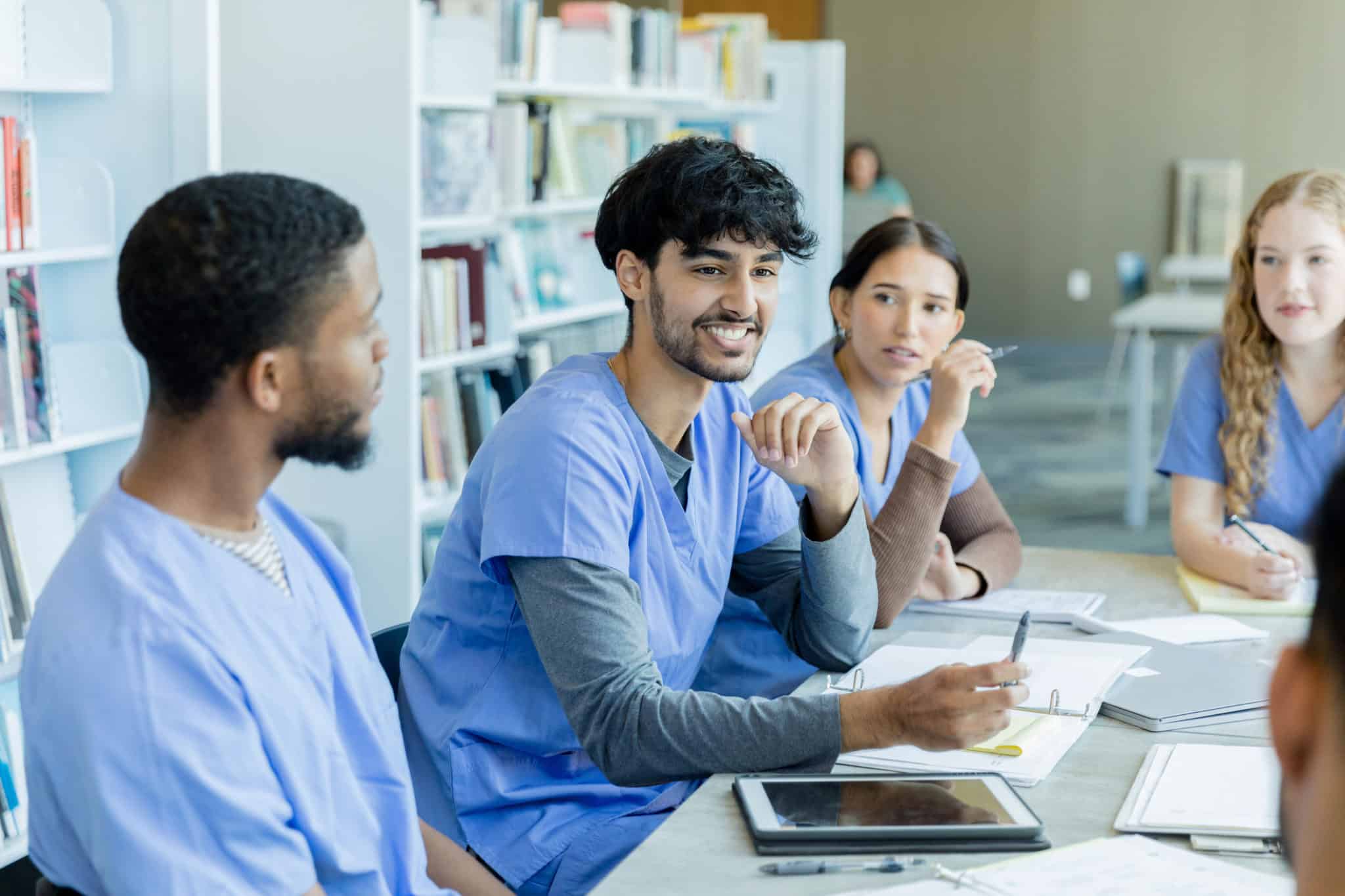 Sitting together at a table in the library, the diverse young adult medical students listen to the discussion between the two young adult men.